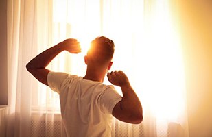 Man waking up refreshed, looking at sunrise through window